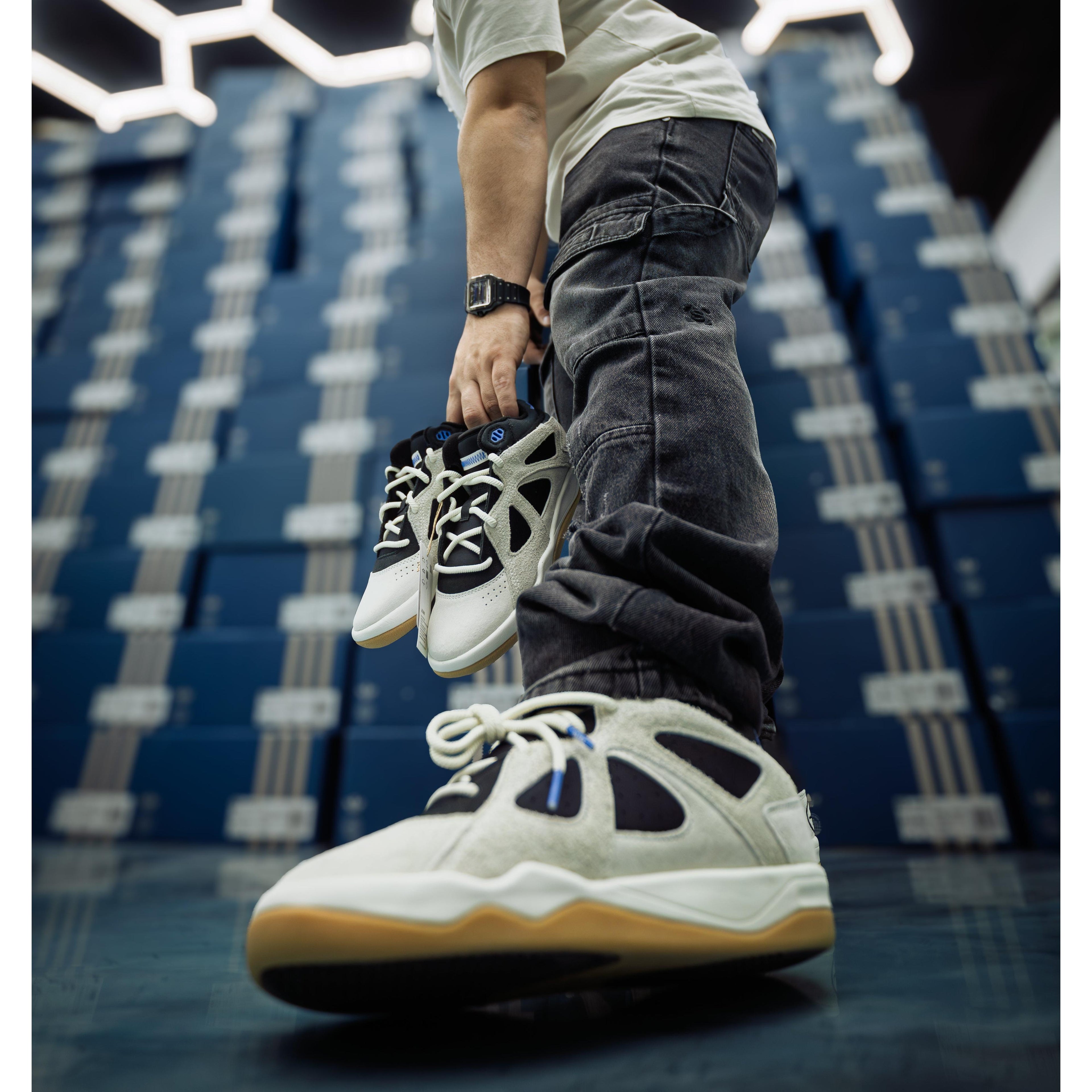 Person holding two small white and black ADIDAS BADBO 1.0 RISE sneakers, wearing a larger pair.