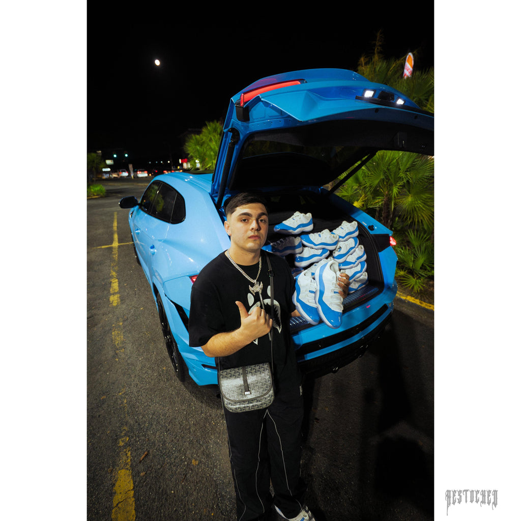 Man holding a Jordan 11 Retro Low University Blue sneaker, with more pairs in a blue car trunk.
