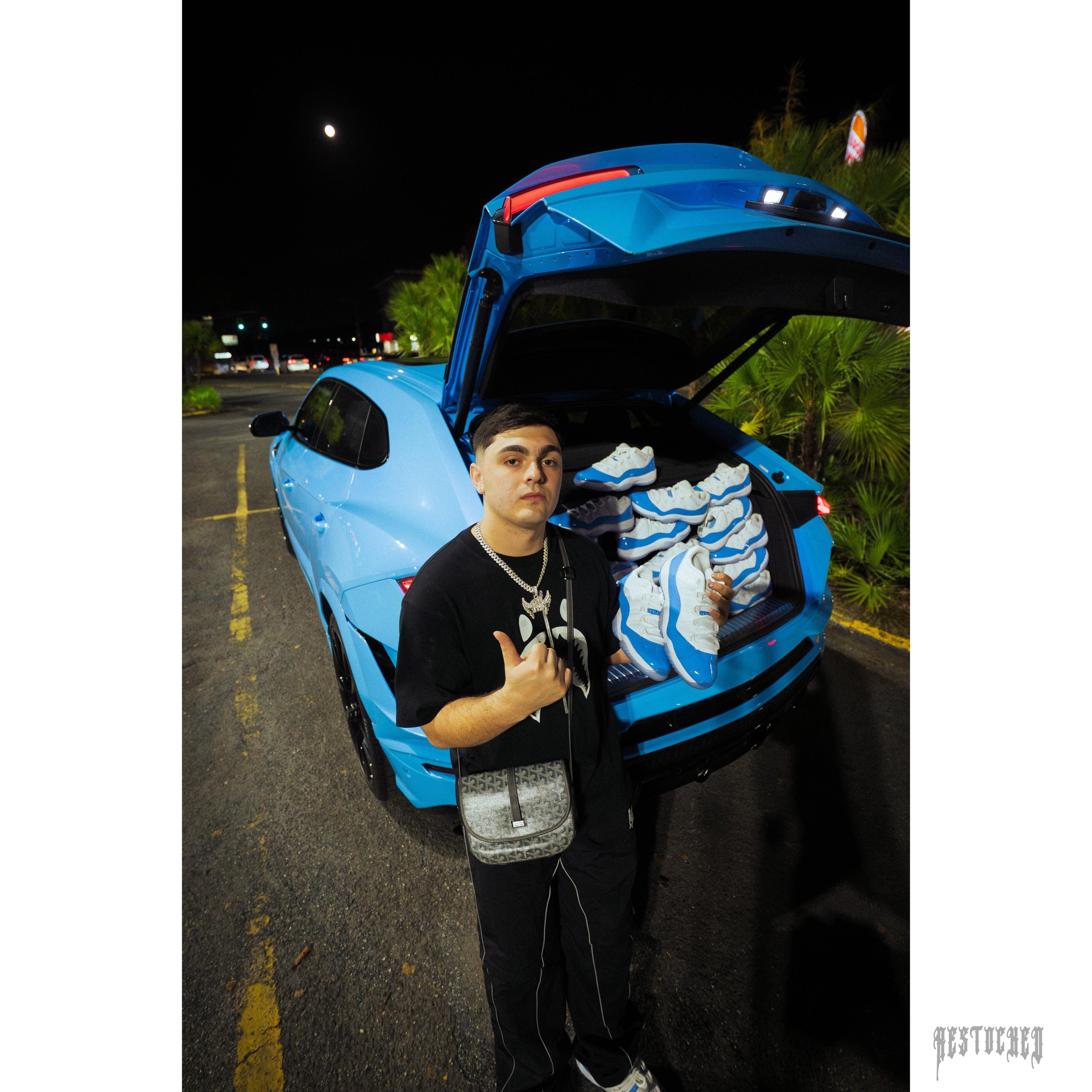Man holding a Jordan 11 Retro Low University Blue sneaker, with more pairs in a blue car trunk.